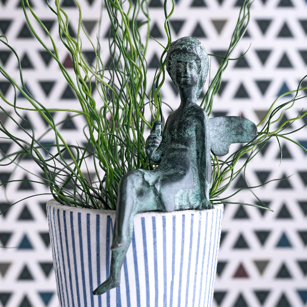 Statue of a child in a striped pot with greenery against a geometric patterned wall.
