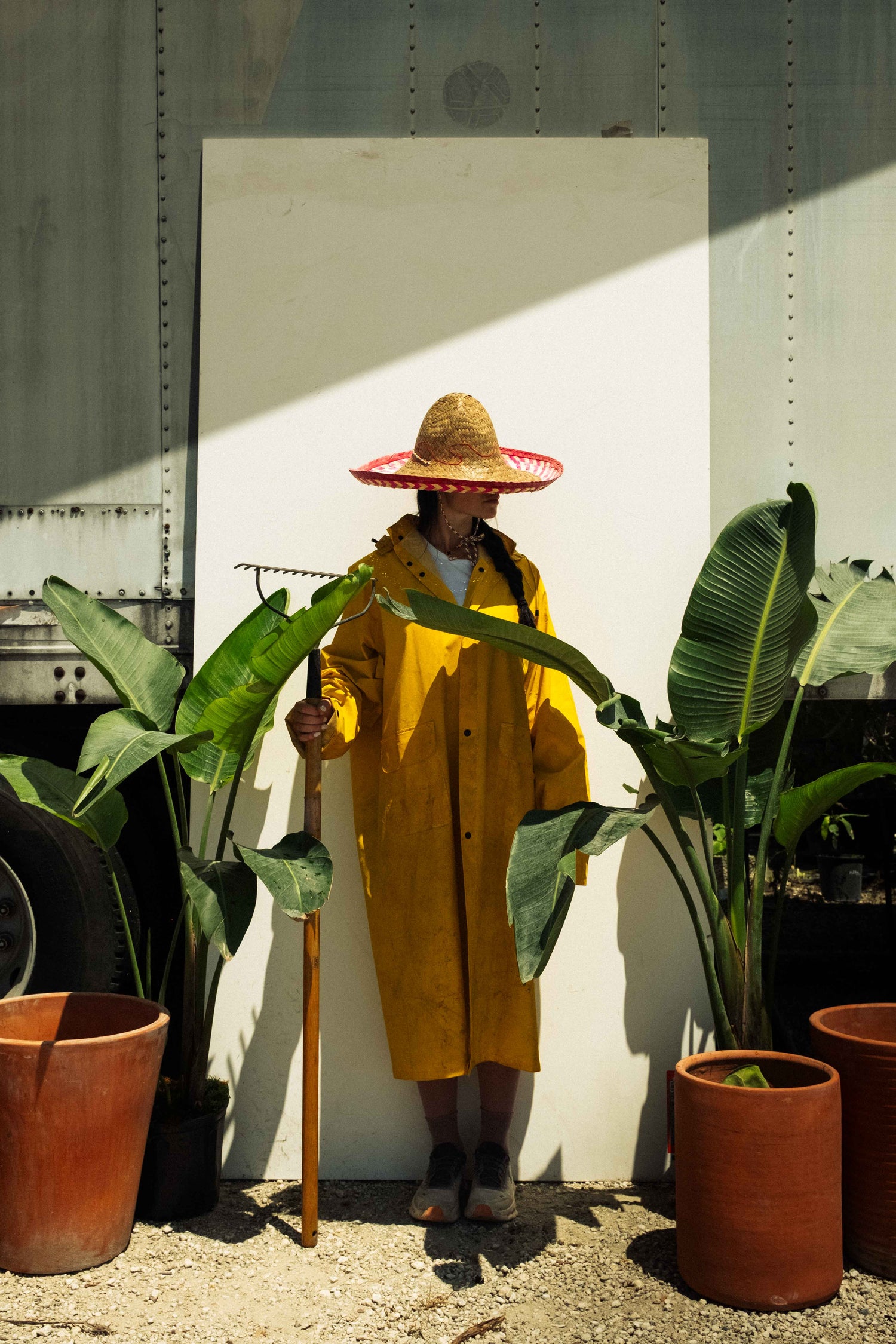 Person in a yellow raincoat and straw hat holding a garden rake among potted tropical plants against a sunlit white backdrop