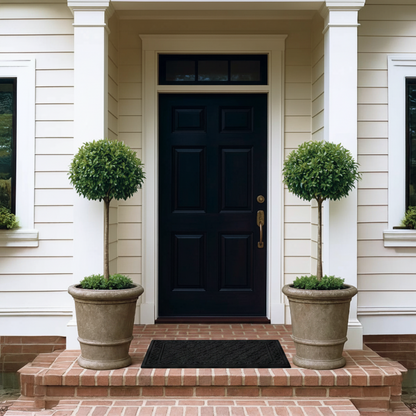 A front porch entryway with a dark navy door centered between white columns, flanked by two large beige planters holding trimmed topiary trees. A black doormat sits on the brick steps below.